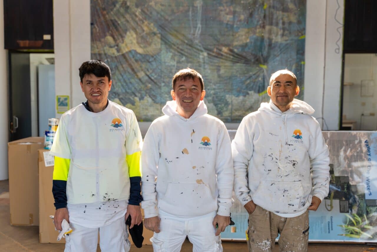 Three men in paint-splattered clothing stand in front of a large map inside a hut. Each wears a white top marked with visible paint stains, suggesting their involvement in a conservation project.