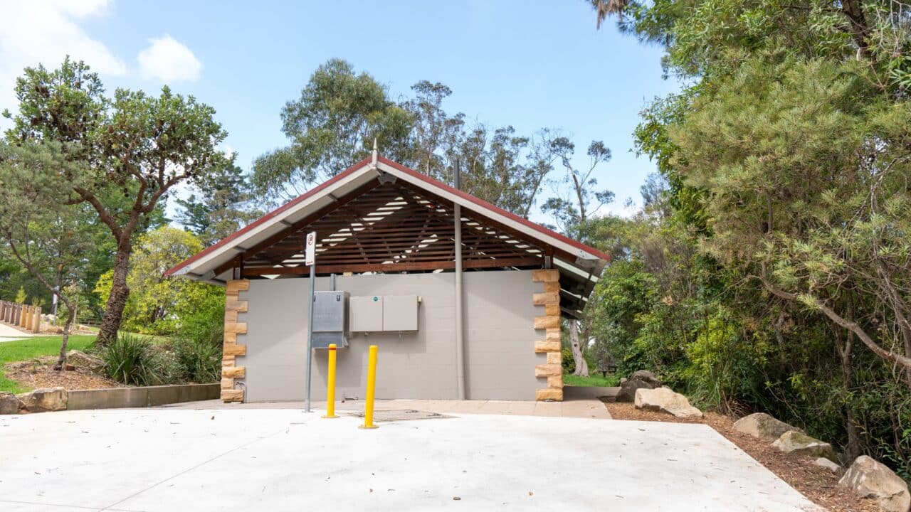 A small public restroom facility with a sloped roof, surrounded by trees and shrubs. Located near Wentworth Falls Lookout, it features two utility boxes and yellow bollards positioned in front of the building, blending functionality with scenic charm.