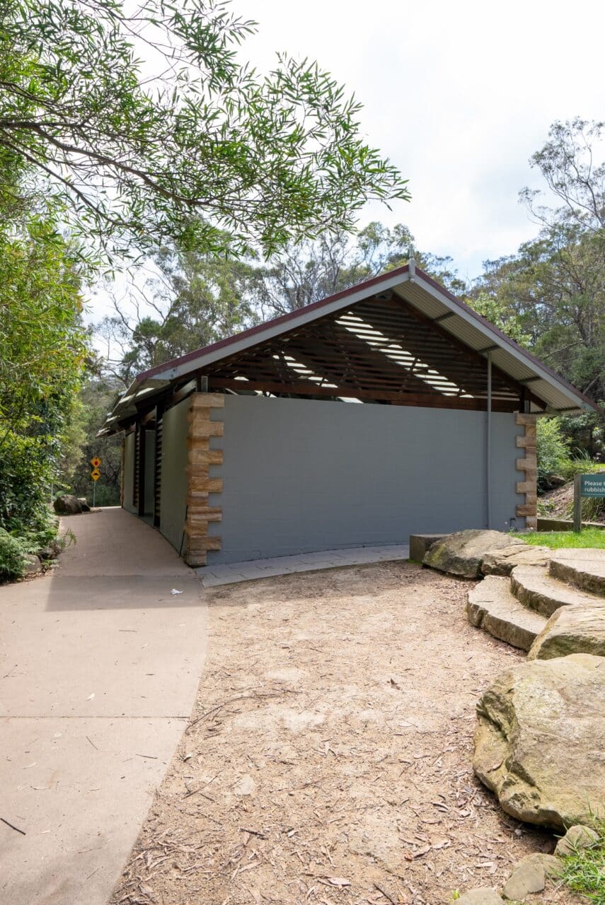 A public restroom building with a green exterior, surrounded by trees and rocks, with a paved pathway leading to the entrance, reminiscent of the serene beauty found at Wentworth Falls Lookout.