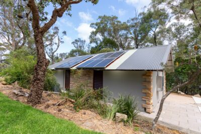 A small building with a corrugated metal roof, featuring solar panels, stands amidst trees and a garden at Wentworth Falls Lookout. This transformed toilet block is accessed by a stone pathway leading to the entrance.