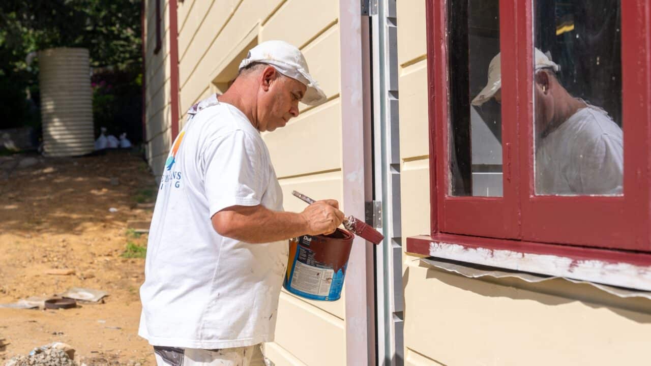 A person in white painter's clothing from a Residential Painting team is painting the edges of a window frame with red paint on the exterior of a cream-colored building.