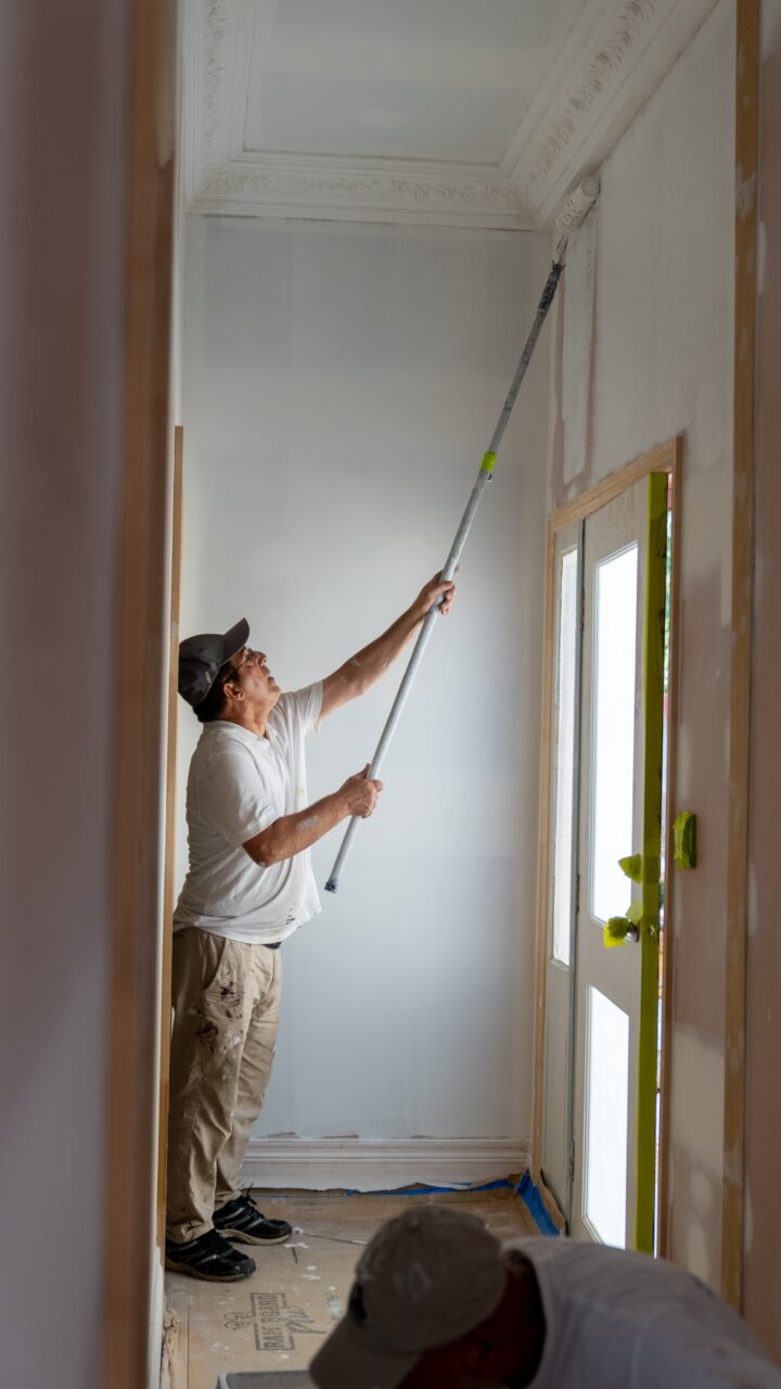 A man in a white shirt and cap delivers professional painting services, using a long-handled roller to paint the top of a wall in a hallway under renovation. Another individual works near the floor, partially visible. This efficient team brings exceptional residential painting to the Blue Mountains area.