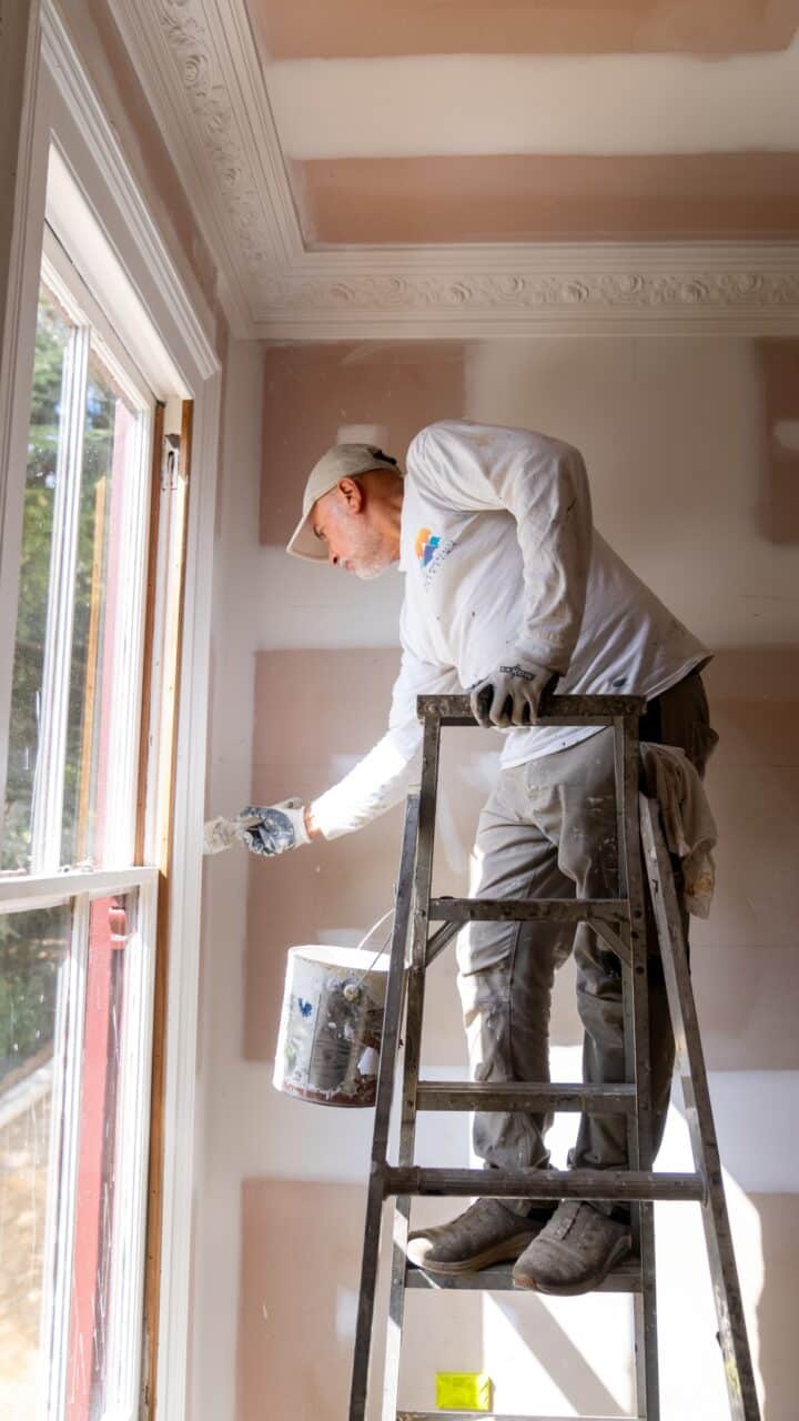 A person standing on a ladder and painting the trim of a window in a room with partially painted walls and a decorative ceiling, showcasing the meticulous craft of residential painting services.