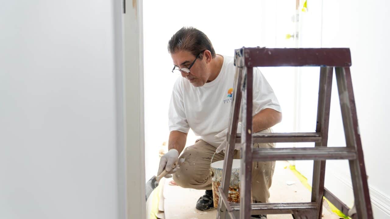 A man in a white shirt and gloves kneels to paint a wall near a stepladder in a brightly lit room, showcasing his residential painting skills. Through the window, the stunning Blue Mountains provide a picturesque backdrop.