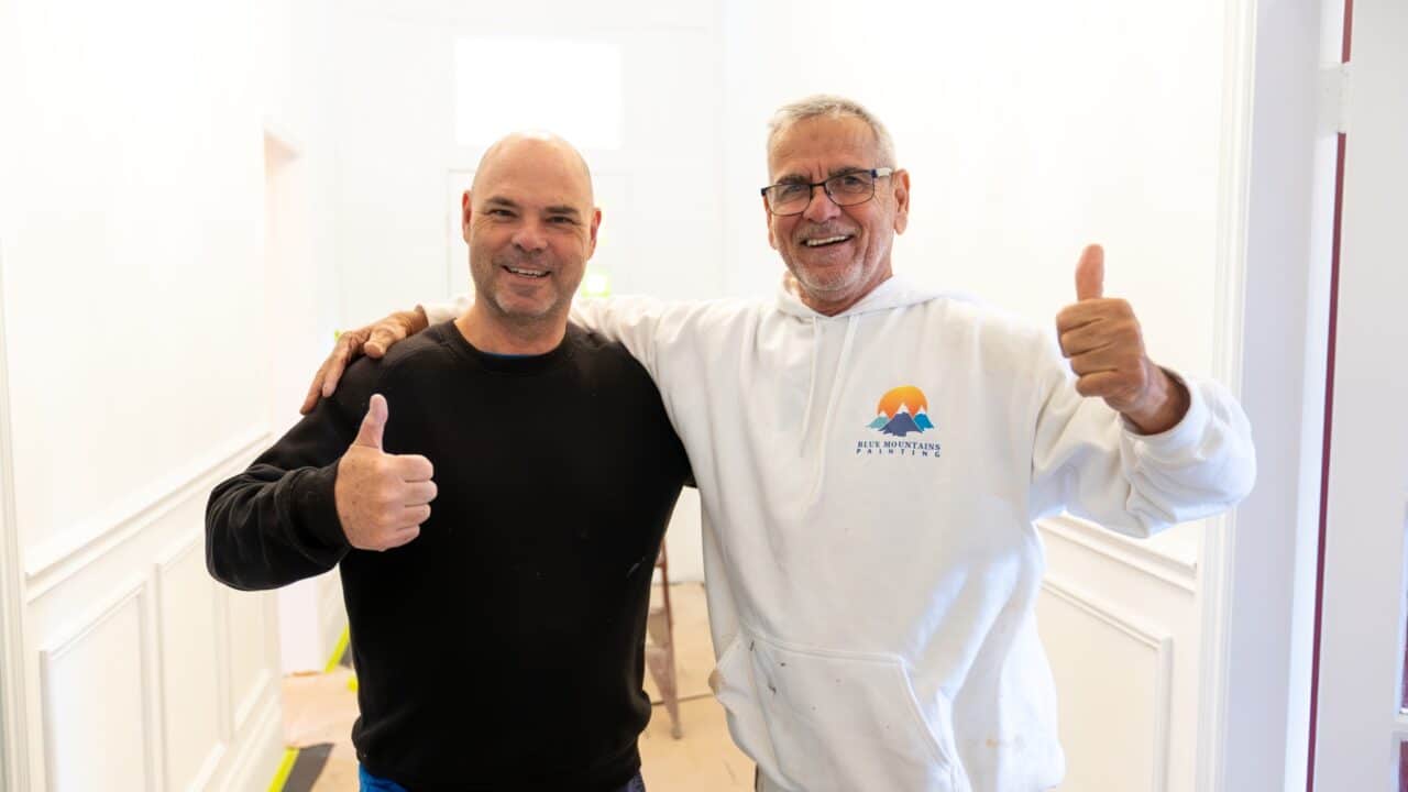 Two men standing side by side in a white-walled room, smiling and giving thumbs up. One man wears a black sweater, and the other sports a white hoodie with a colorful logo. Their happy expressions suggest another successful residential painting project in the Blue Mountains.