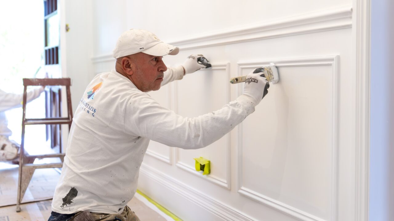 A man wearing a cap and gloves paints decorative molding on a white wall, showcasing the precision of Residential Painting Services. A ladder and other painting supplies are in the background, hinting at a meticulous project set in the serene Blue Mountains.