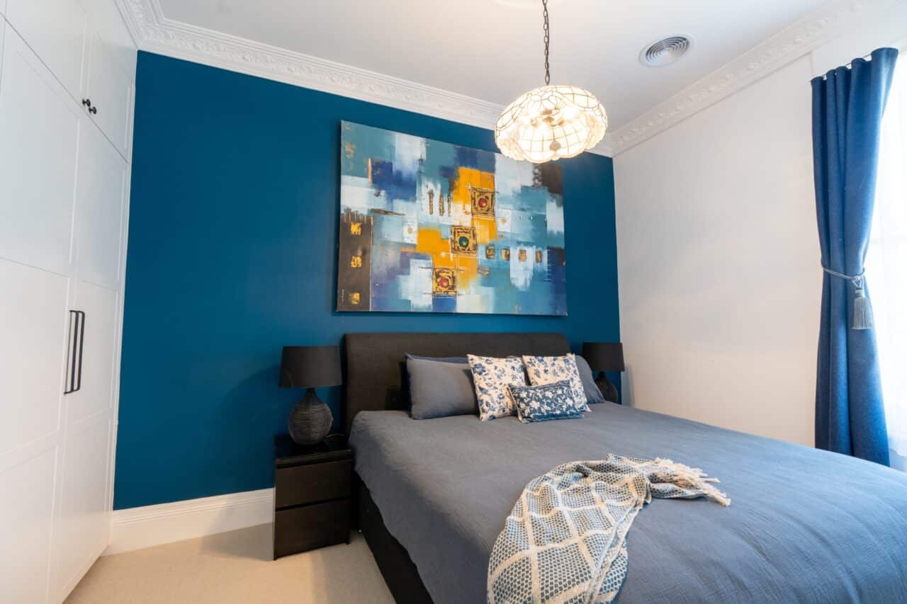 A bedroom with a blue accent wall reminiscent of the Blue Mountains, abstract painting, dark bedframe, blue bedding, blue curtains, white closet doors, and two black nightstands with matching lamps. A round ceiling light fixture is overhead.