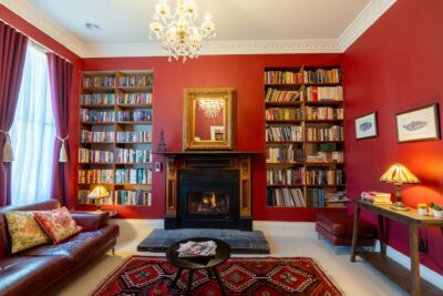 A cozy living room with red walls, built-in bookshelves, a fireplace with a mirror above it, a chandelier, a leather sofa, a patterned rug, two lamps, and a window with red curtains showcases the expert interior design services inspired by the serene Blue Mountains.