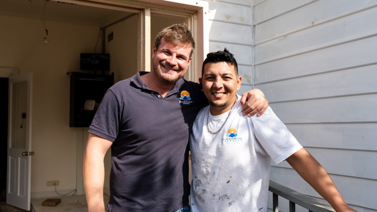 Two men smiling with arms around each other, standing outside a white building. Both wearing shirts with a logo.