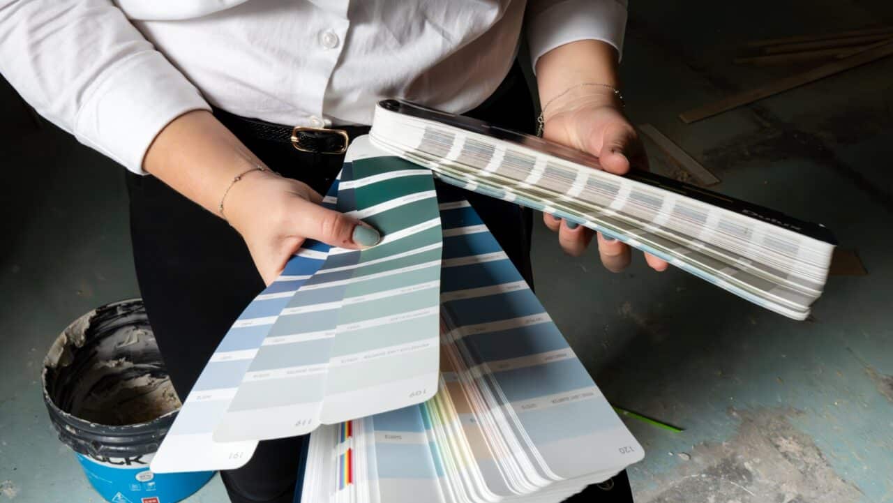 A person holding several color sample swatches, likely for paint, inside a room under renovation. A paint bucket and construction materials are visible in the background, showcasing expert interior design decisions in progress.