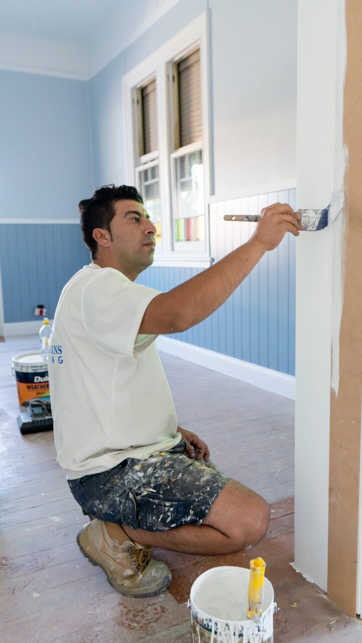 A man wears a white T-shirt and denim shorts, kneels on the floor, and paints a white wall with a brush in a room with blue wainscoting, showcasing his expert Interior Painting Services.