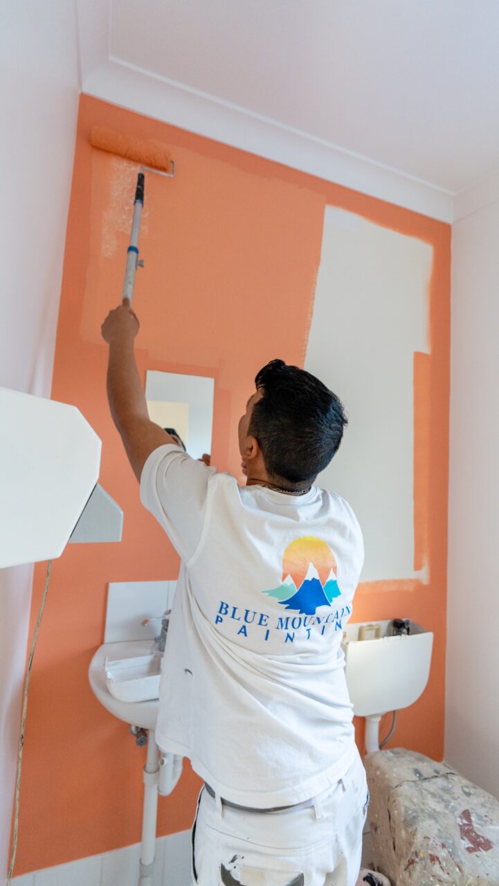 A person in a white T-shirt with a "Blue Mountain Painters" logo is painting a wall orange with a roller in a room that has a sink and small basin, showcasing the impeccable Strata Painting Services provided across the Blue Mountains.