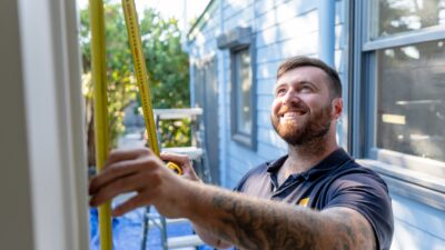 A man in a polo shirt with tattoos on his arms measures a wall outdoors using a measuring tape. He stands in front of a light blue house, smiling while taking the measurements, showcasing the precision of Expert Carpentry Services amidst the serene backdrop of the Blue Mountains.