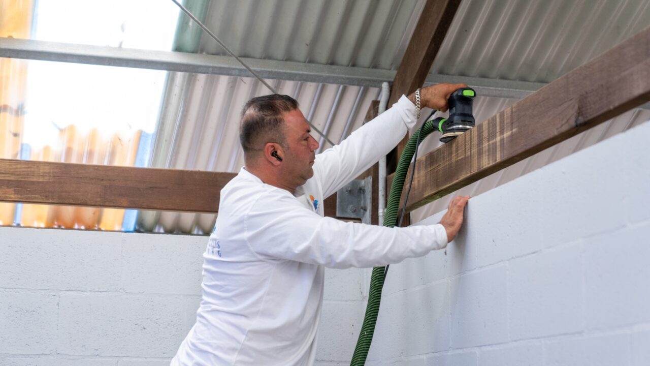 A person in a white long-sleeve shirt sands a piece of wood with a power sander inside a building, likely preparing materials for the Wentworth Falls Lookout transformation.