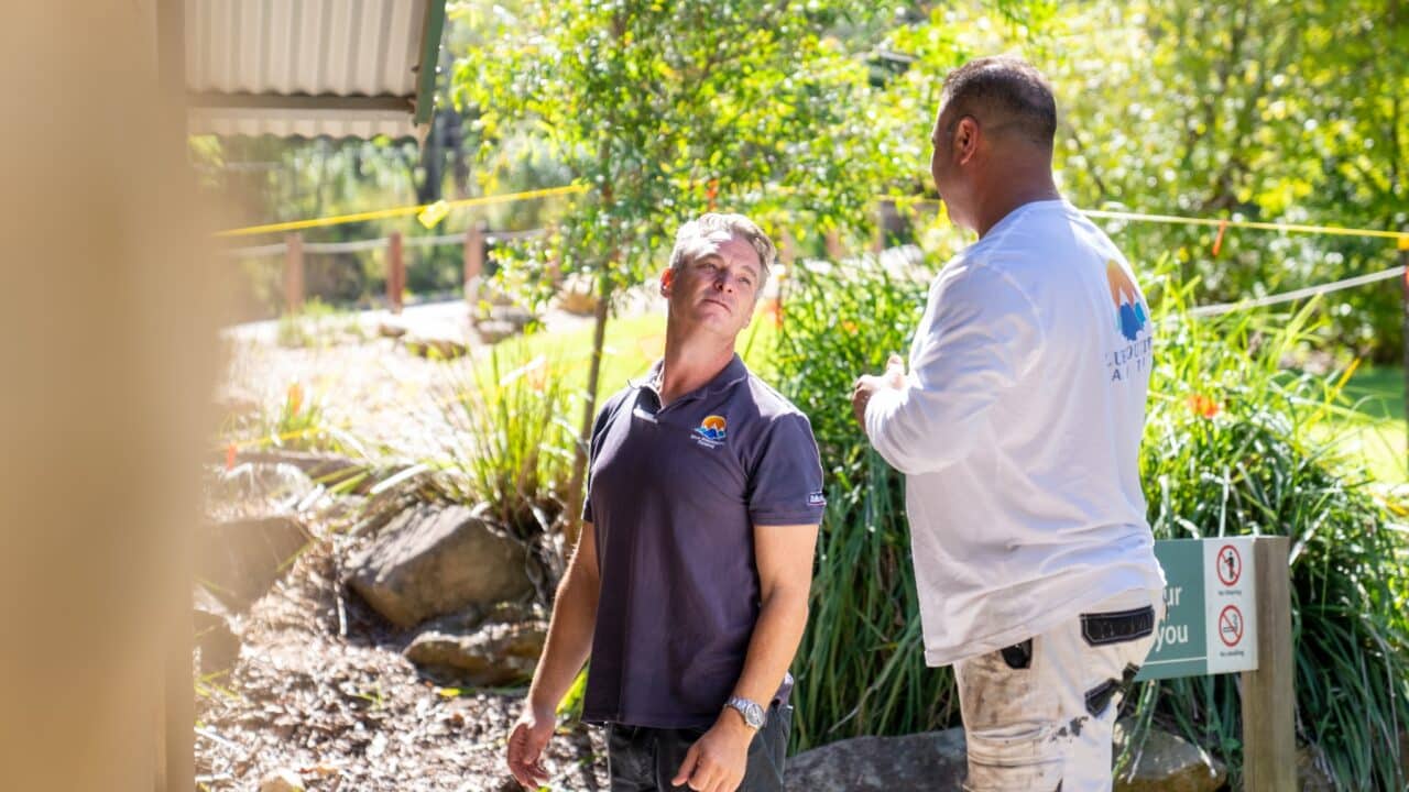 Two men in casual work attire are conversing outdoors at Wentworth Falls Lookout, surrounded by greenery and trees. One man wears a white shirt and the other a dark shirt; both exhibit attentive body language, possibly discussing their Project on the Toilet Block Transformation.