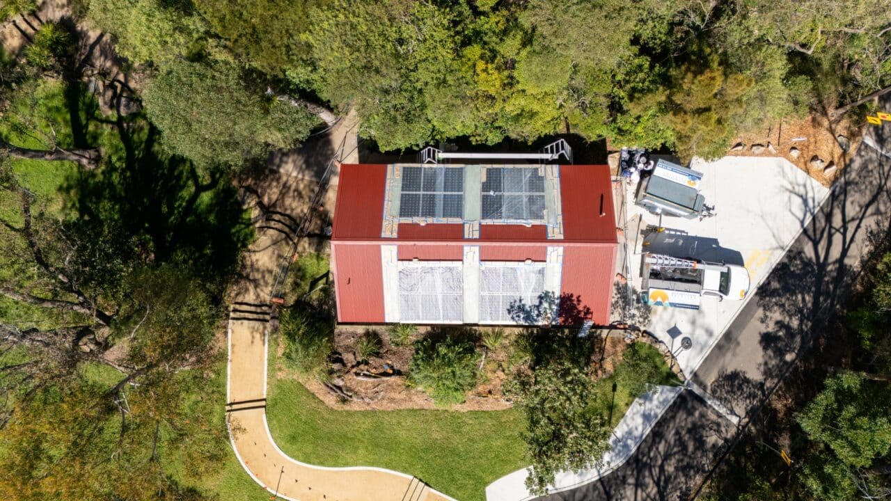 Aerial view of a red-roofed building surrounded by trees with a parking area and a vehicle parked nearby. Pathways and landscaping are visible in the vicinity, showcasing the Wentworth Falls Lookout transformation project. Solar panels are installed on the roof.