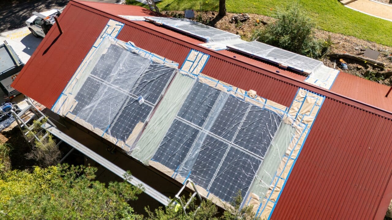 Aerial view of a red rooftop with partially installed solar panels covered in protective plastic sheeting, surrounded by greenery and a ladder leaning against the side; part of the exciting Toilet Block Transformation project near Wentworth Falls.