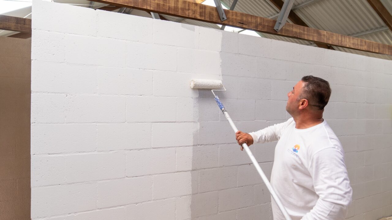 A man in a white shirt is transforming a cinder block wall using a long-handled roller, painting it white in an indoor setting with a metal roof. This scene feels reminiscent of the fresh renovations seen at Wentworth Falls Lookout.