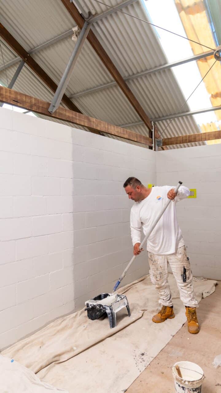 A person in white attire and brown boots is painting a white wall with a roller in a room with a corrugated metal roof. The project, aimed at the Wentworth Falls Lookout toilet block transformation, includes painting supplies neatly arranged on a drop cloth on the floor.