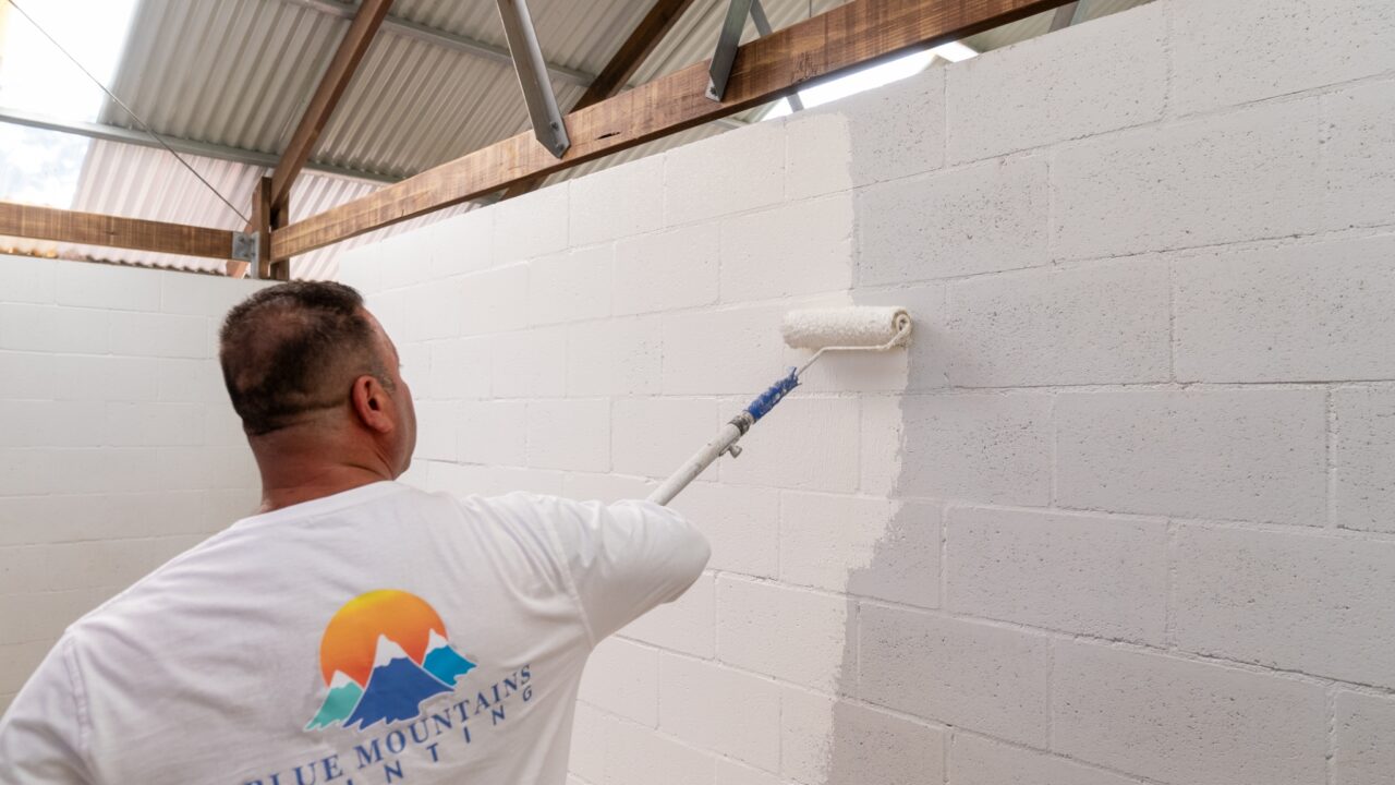 A person is painting a white concrete wall with a roller attached to an extension pole. The individual is wearing a white shirt with a colorful logo featuring mountains and the text "BLUE MOUNTAINS PAINTING." This project marks the start of the Toilet Block Transformation at Wentworth Falls Lookout.