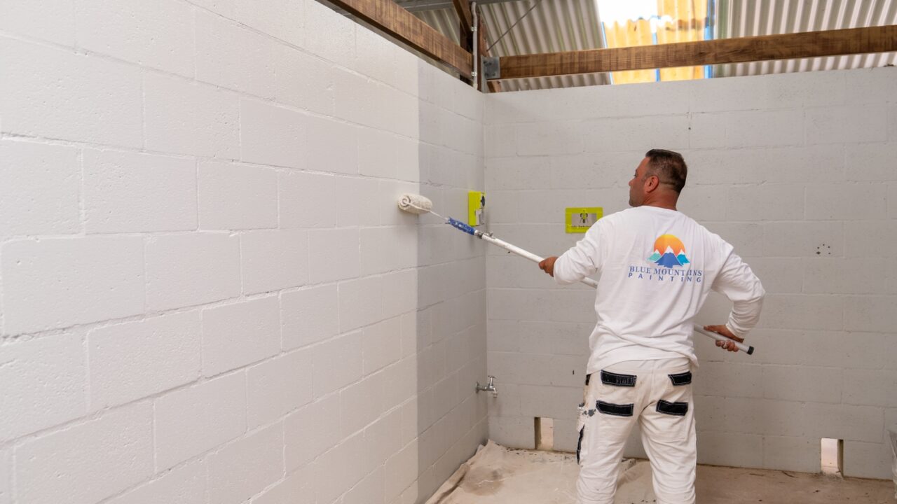 A man in a white shirt and pants is painting a cinder block wall with a roller, part of the Toilet Block Transformation at the Wentworth Falls Lookout Project.