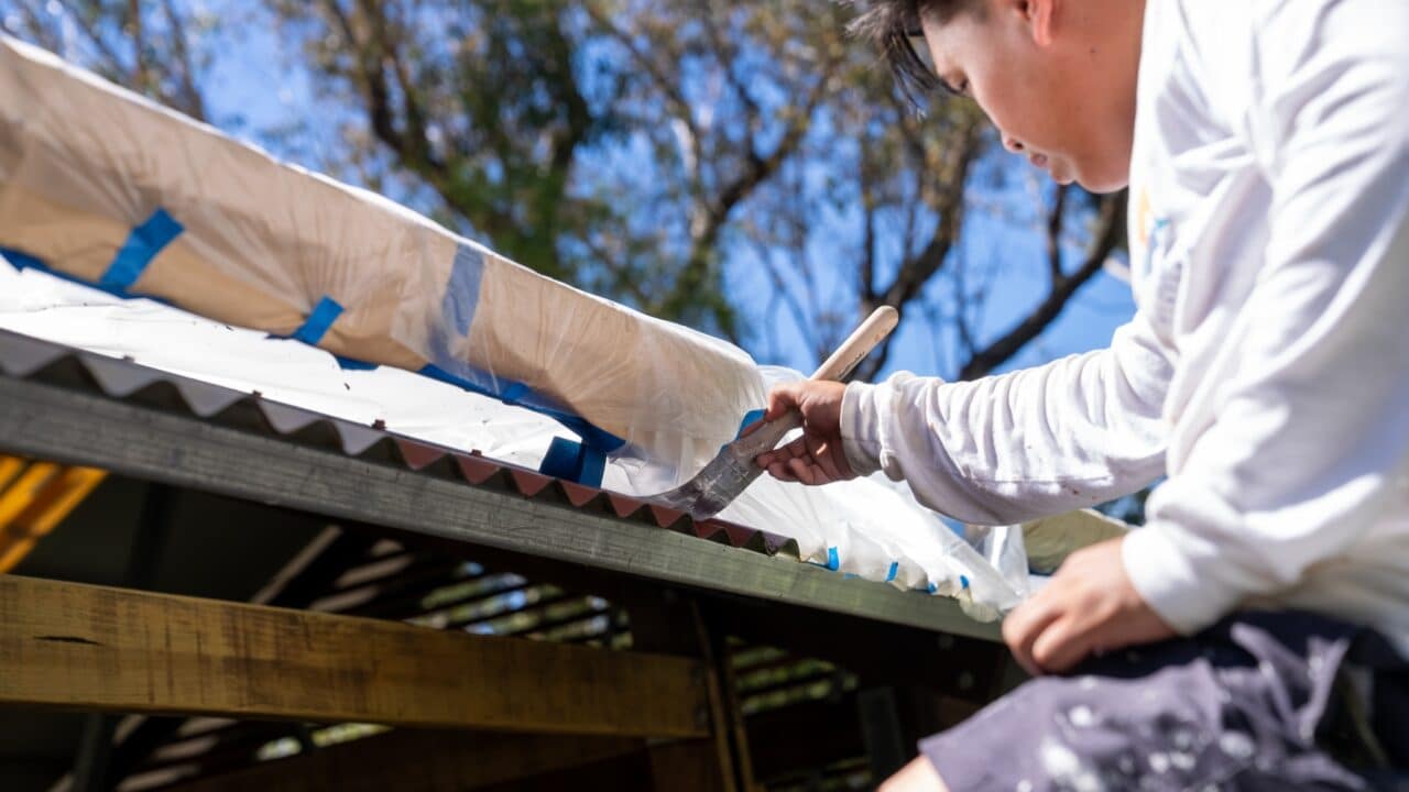 A person is painting corrugated metal roofing while perched on a ladder, working on the Toilet Block Transformation at Wentworth Falls Lookout. The roof is partially covered with protective plastic, and trees and a clear sky serve as the picturesque backdrop.