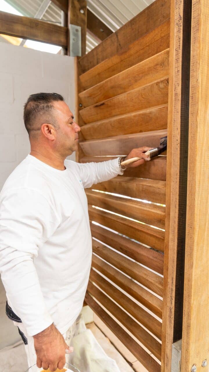 A person in a white shirt and paint-stained pants is applying a dark stain to a wooden fence using a brush in an outdoor setting, as part of the Wentworth Falls Lookout Project.