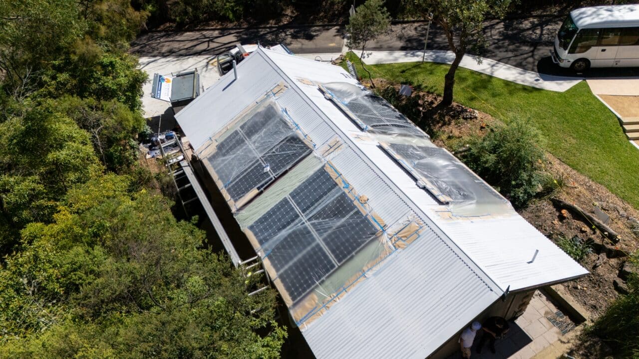 Aerial view of a house with solar panels installed on the roof, partially covered by transparent plastic, surrounded by trees and near the Wentworth Falls Lookout, with a road and a parked bus visible in the background.