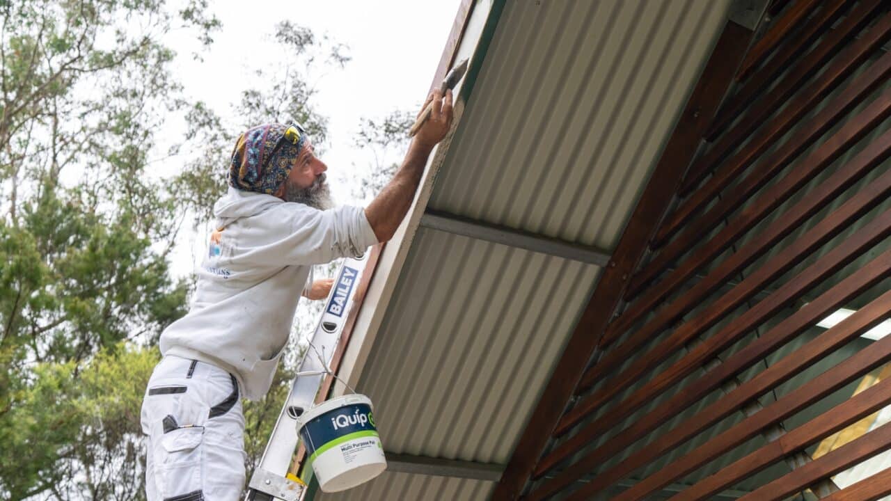 A person wearing white clothing and a colorful headband is standing on a ladder, painting the fascia of a house roof with a roller brush as part of the Lookout Project. A paint bucket is attached to the ladder. Trees are in the background.