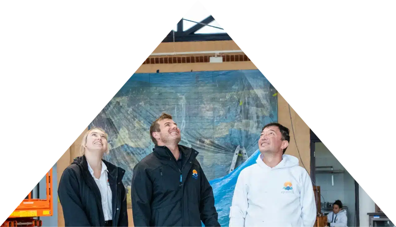 Three people in jackets stand indoors, looking up with smiles, in front of a mural.