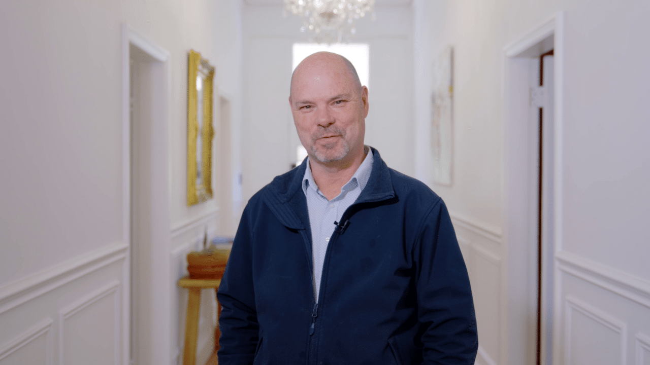 A man with a bald head and a short beard, wearing a blue jacket, stands in the well-lit hallway of Heritage House, adorned with white walls and a chandelier overhead.