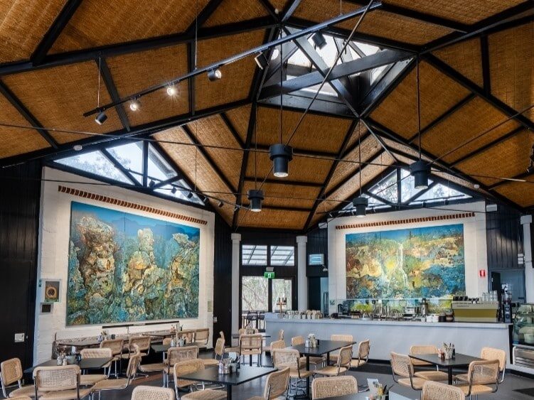 A spacious, modern café with wicker chairs and black tables. The ceiling features a geometric pattern reminiscent of architectural conservation efforts, and large colorful murals adorn the walls. The café counter is seen in the background, giving a cozy hut-like feel to the space.