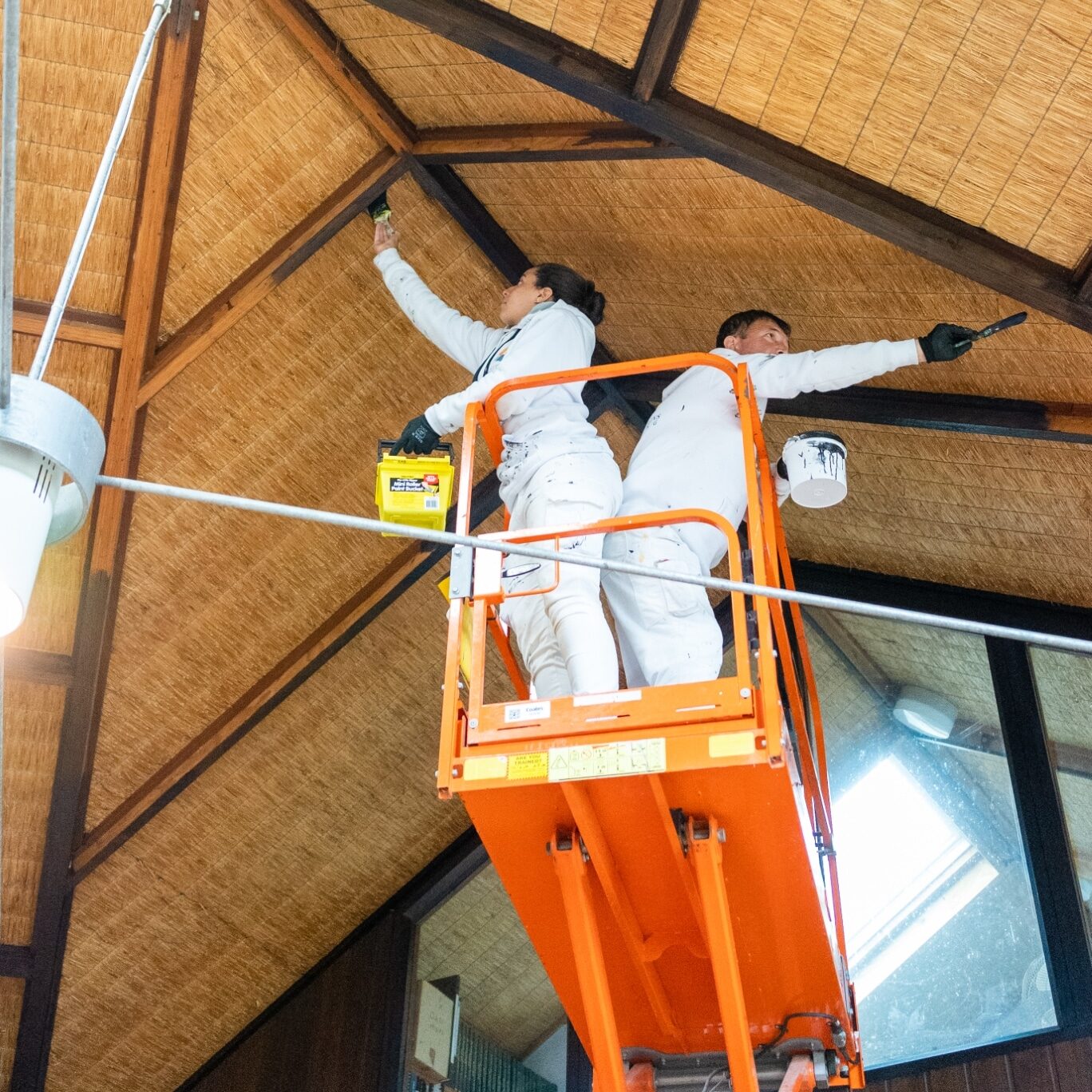 Two individuals in white overalls on an orange scissor lift are painting the wooden ceiling of a high room. One holds a paint can while the other applies paint with a brush, showcasing their expertise as part of Commercial Painting Services in the Blue Mountains.