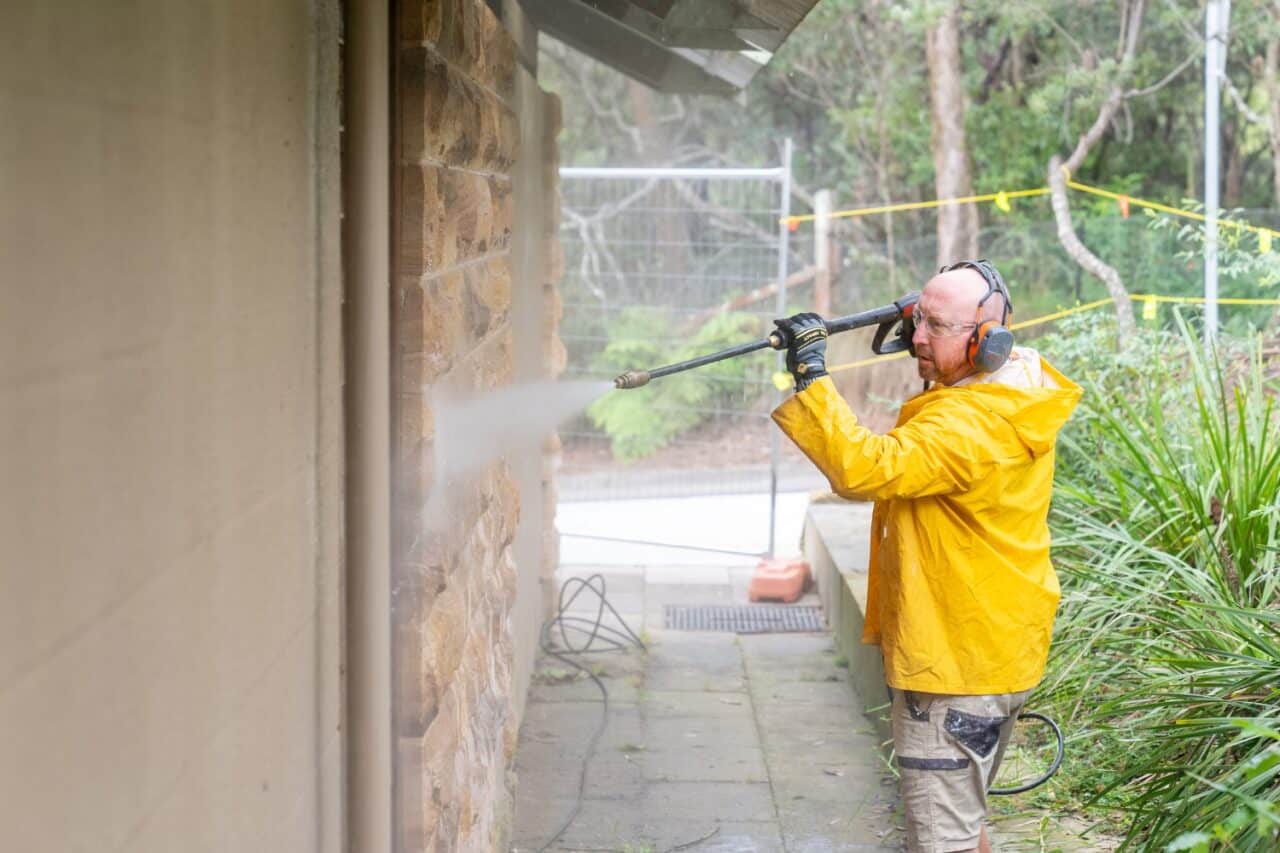 a man in a yellow raincoat pressure cleans a stone wall outdoors, showcasing the power cleaning of hard surfaces. equipped with ear protection and gloves, he navigates around plants and temporary fencing with precision.